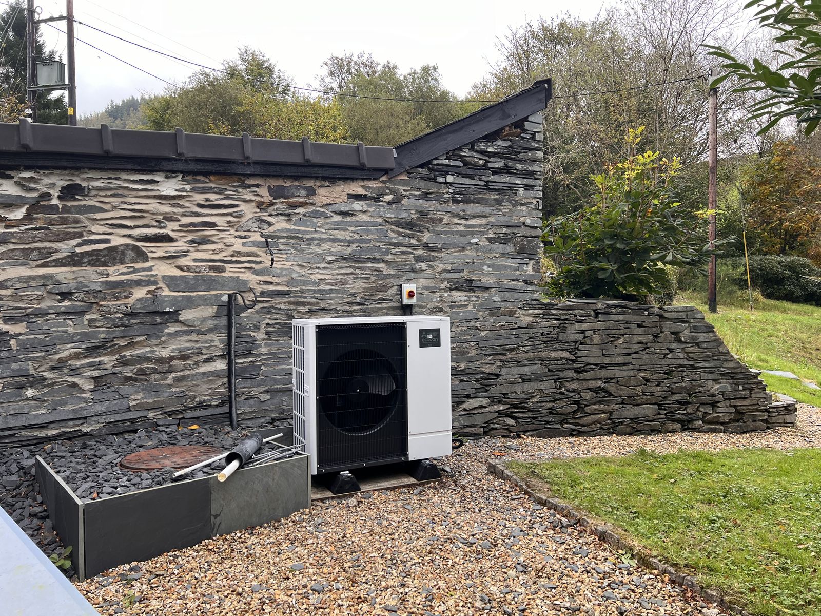 White Mitsubishi Ecodan air source heat pump on a gravel base in front of an old slate stone wall